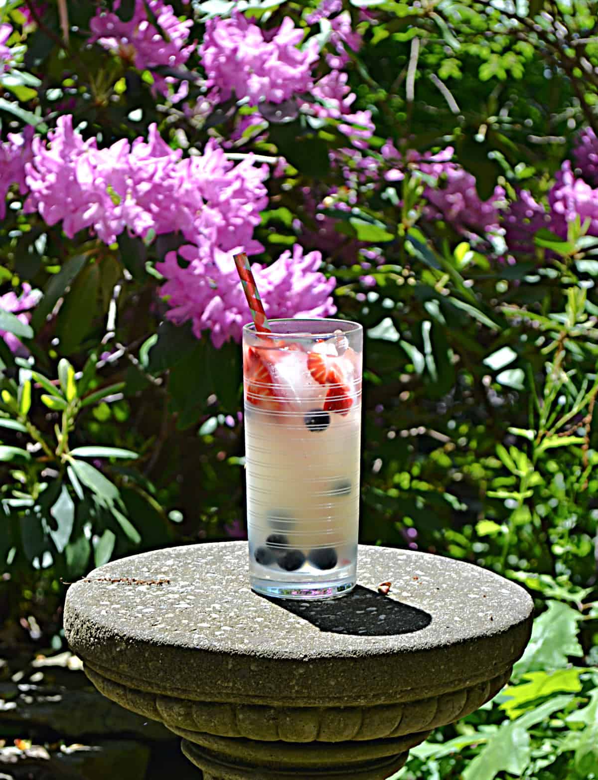 A glass of lemonade on a stone pedestal with pink flowers behind the glass.