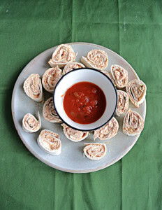 A plate of pinwheels with a bowl of salsa in the middle.