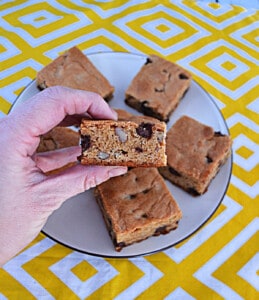 A hand holding a chocolate chip blondie with a plate of blondies behind it.
