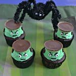 Four Frankenstein Cupcakes with green frosting, edible eyes, and a peanut butter cup hat sitting on a green striped background with a huge spider towering over them in the background.