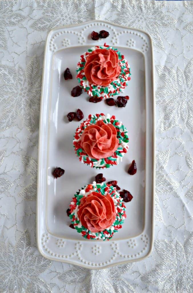 A white platter with three cupcakes on it. Each cupcake has a white ring of frosting with sprinkles on it with red frosting piped in the middle on a white background. 