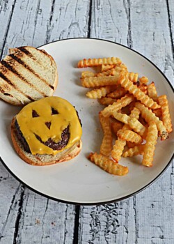 A plate with a piece of cheese that looks like a Jack-o-lantern face on a toasted bun along side a pile of fries.