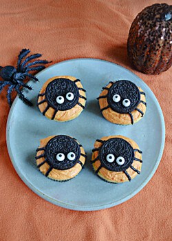 A plate of cupcakes with Oreo spiders, a toy spider on the plate, and a pumpkin behind the plate.