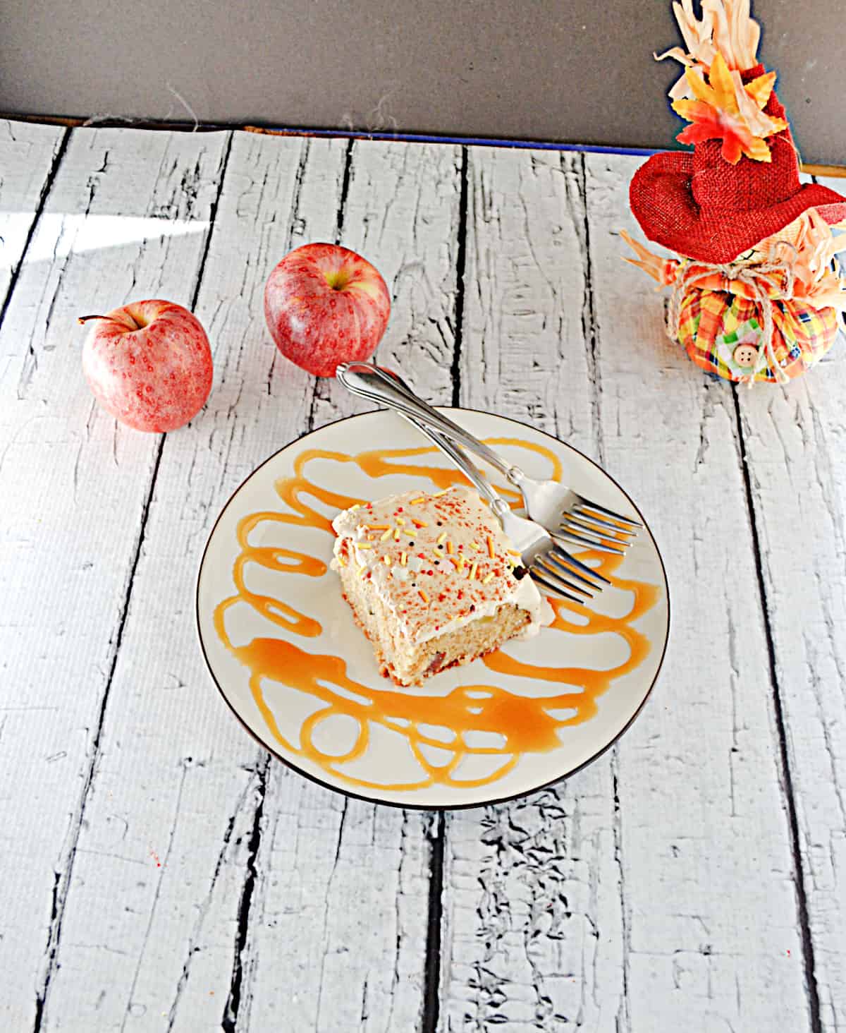 A top view of a slice of Apple Pie Cake with two apples beside it, a fork on the plate, and a drizzle of caramel sauce. 