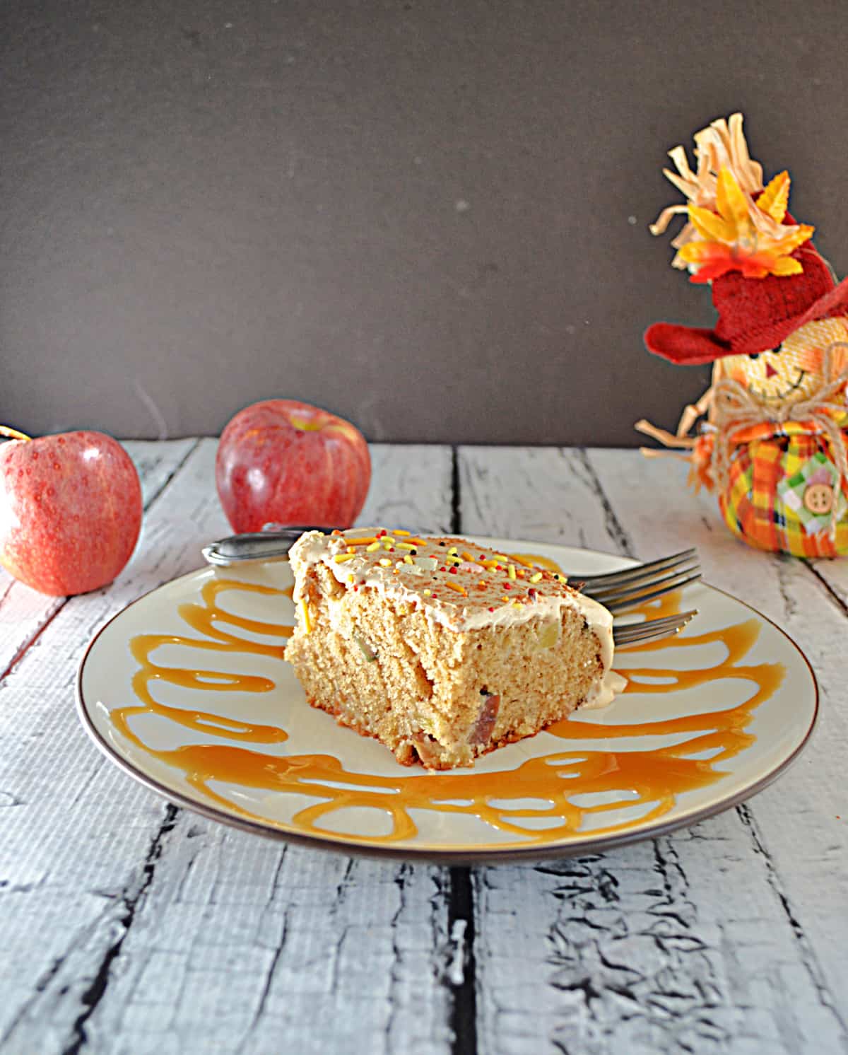 A caramel drizzled plate with a slice of apple cake on it and 2 apples and a scarecrow in the background.