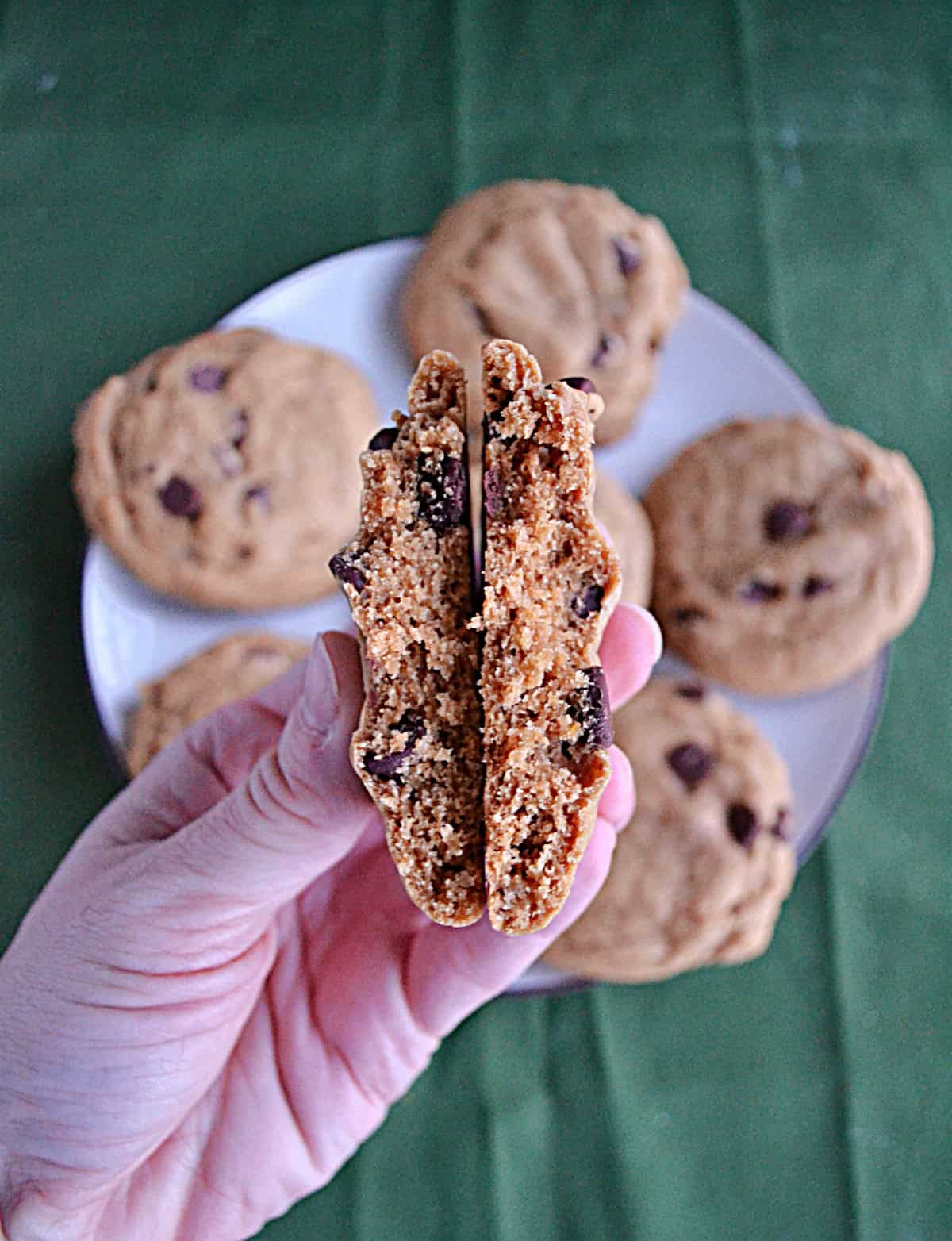 A hand holding a chocolate chip cookie broken in half with a plate of cookies in the background.