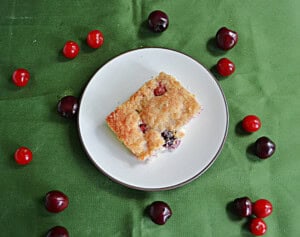 A plate with a piece of cherry coffee cake on it and fresh cherries surrounding the plate.