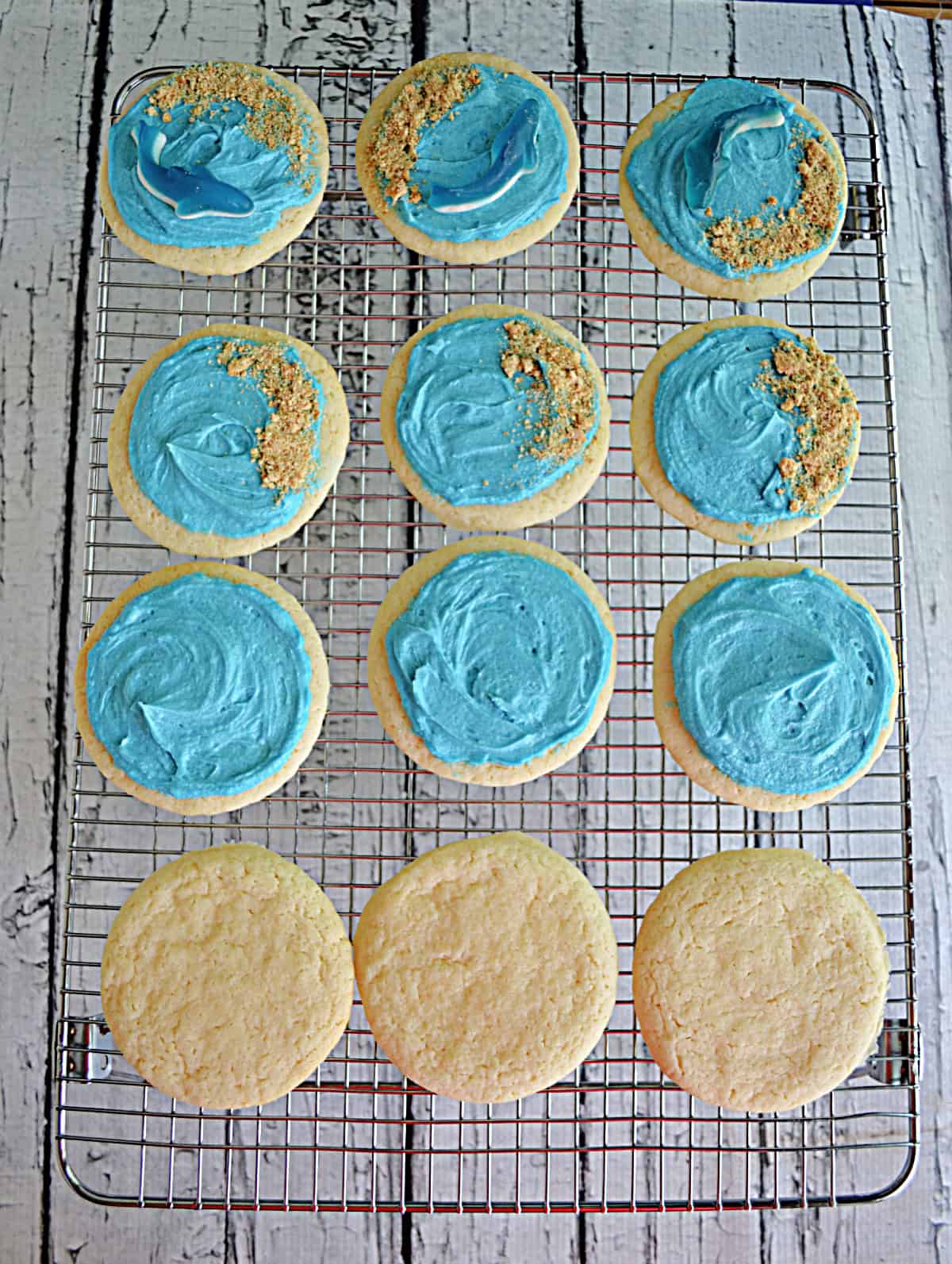 A wire rack with a dozen cookies on it. Three have sharks on them, three have frosting, three are plain, and three have frosting and graham cracker crumbs.