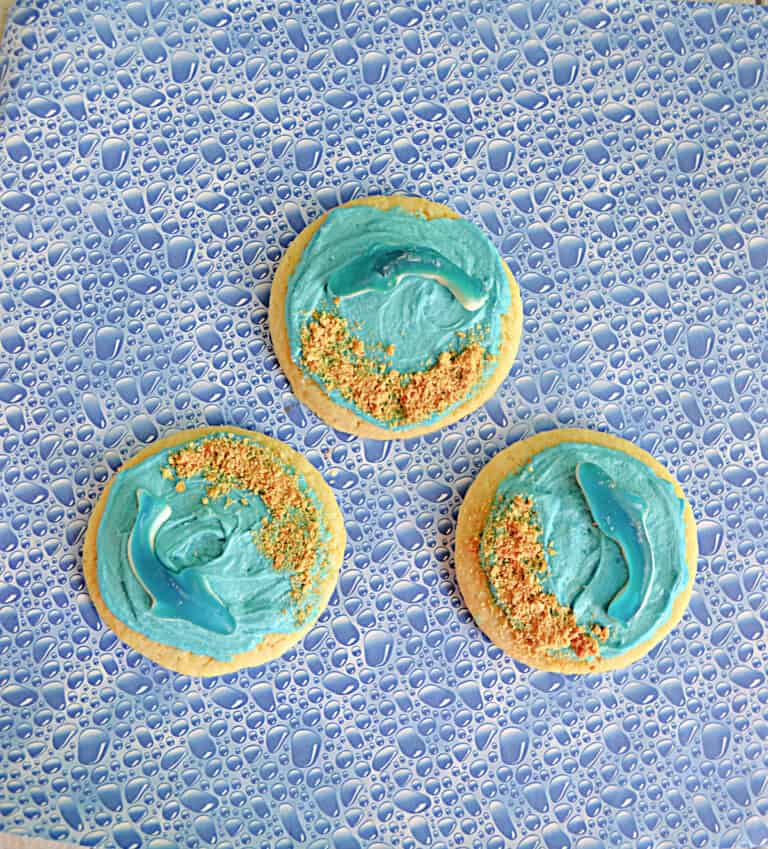 Three sugar cookies with blue frosting, graham cracker crumbs, and gummy sharks on each.