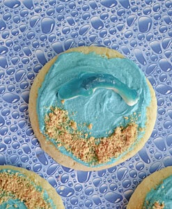 A close up of a sugar cookie with blue frosting, graham cracker sand, and a blue shark gummy.