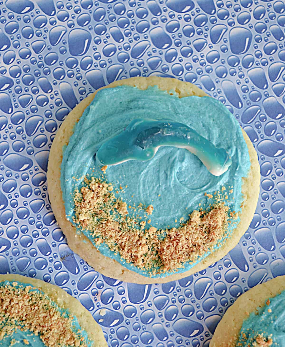 A close up of a sugar cookie with blue frosting, graham cracker sand, and a blue shark gummy.