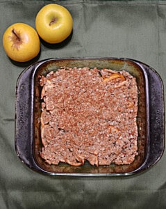 A baking dish of apple crisp with two apples behind the dish.